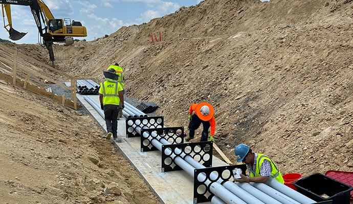 Workers are installing underground conduit pipes in a trench at a construction site.