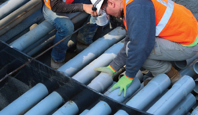 Two construction workers installing and aligning gray utility conduits in a duct bank frame.